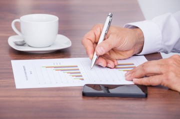 Businessman Writing On Document With Cup And Cellphone On Desk